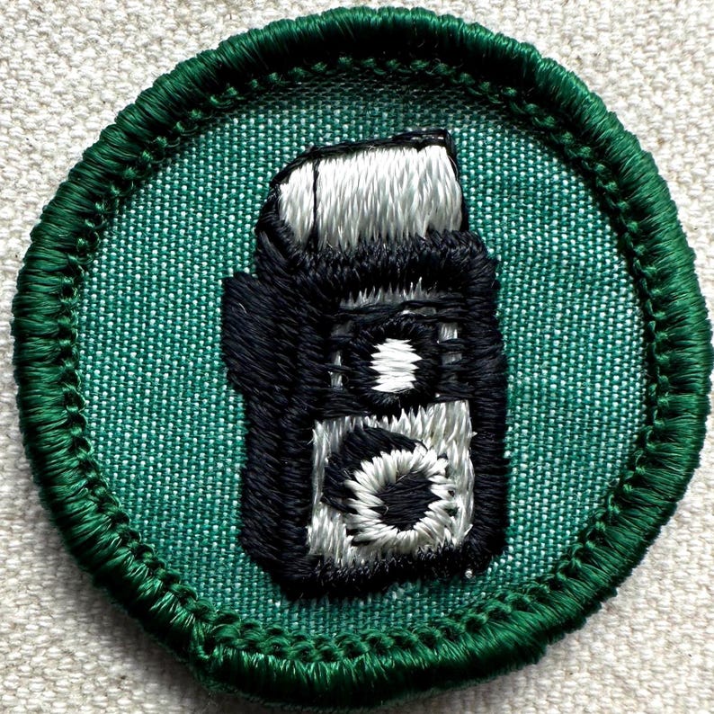 May include: Embroidered patch featuring a black and white camera design on a teal background. The patch has a green border and is likely for a scout or hobbyist. The camera design is detailed with white and black thread.