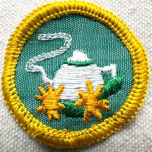 May include: Embroidered patch featuring a white teapot with steam rising, set against a green background. The design includes yellow starbursts and green leaves, all framed by a yellow border.