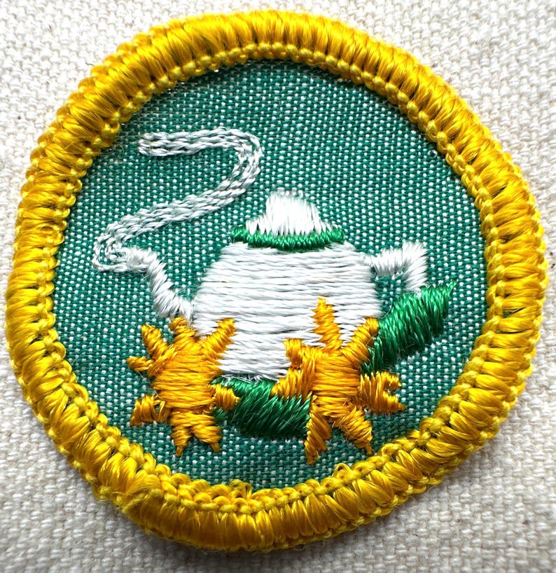 May include: Embroidered patch featuring a white teapot with steam rising, set against a green background. The design includes yellow starbursts and green leaves, all framed by a yellow border.