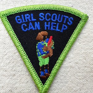 Puede incluir: Un parche triangular de las Girl Scouts con fondo negro y borde verde. El texto "GIRL SCOUTS CAN HELP" est&aacute; bordado en azul. Una figura bordada en azul, verde y marr&oacute;n sostiene un objeto rojo.