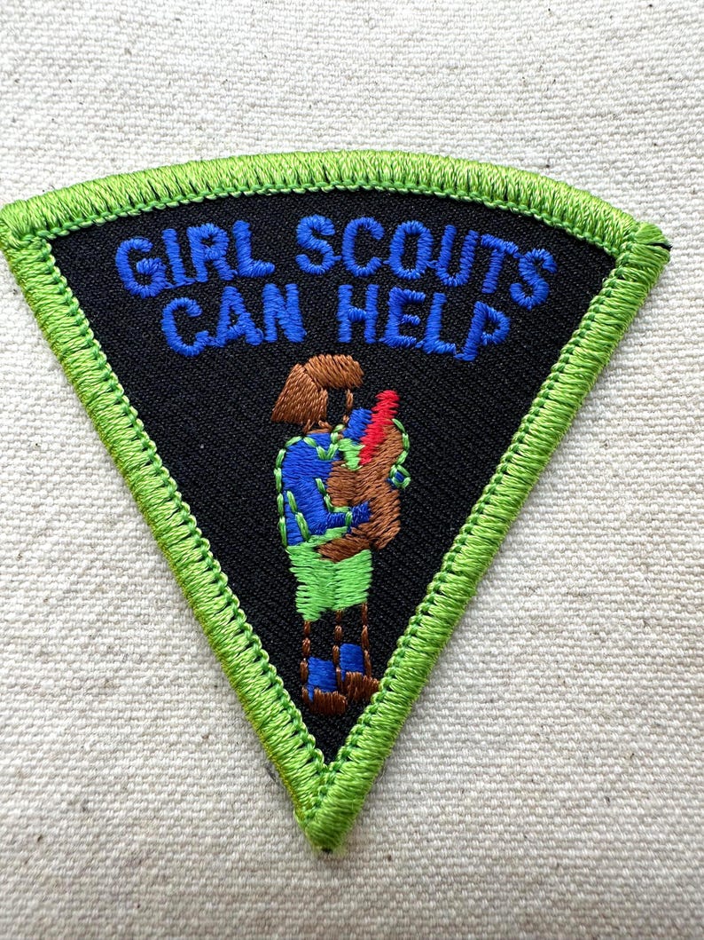 Puede incluir: Un parche triangular de las Girl Scouts con fondo negro y borde verde. El texto "GIRL SCOUTS CAN HELP" est&aacute; bordado en azul. Una figura bordada en azul, verde y marr&oacute;n sostiene un objeto rojo.