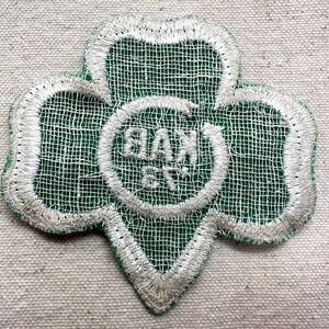 May include: A green and white embroidered patch in the shape of a three-leaf clover. The patch features the text "BAX 6TH" in a white circle at the center. The edges are outlined in white thread.