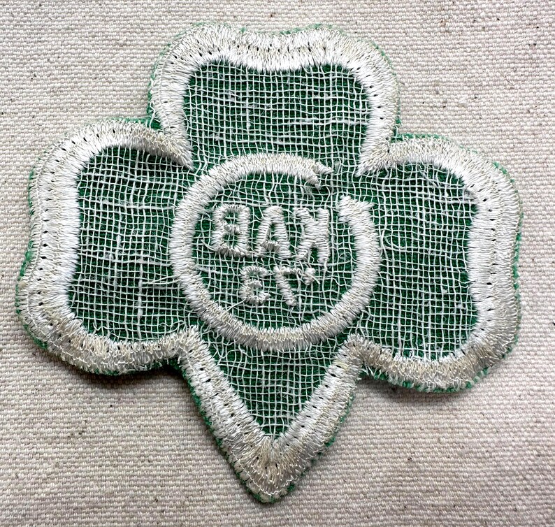 May include: A green and white embroidered patch in the shape of a three-leaf clover. The patch features the text "BAX 6TH" in a white circle at the center. The edges are outlined in white thread.