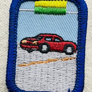 May include: Embroidered patch featuring a red car on a gray road, set against a light blue sky. The patch has a blue border and a green and yellow design at the top. The car has black tires and a detailed design.