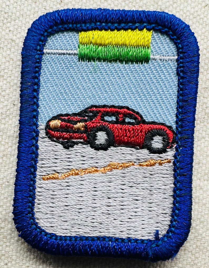 May include: Embroidered patch featuring a red car on a gray road, set against a light blue sky. The patch has a blue border and a green and yellow design at the top. The car has black tires and a detailed design.
