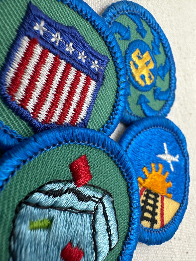 May include: Close-up of several embroidered patches with blue, green, red, white, and yellow details. One patch features a design of the American flag. Other patches have various designs, including a plane, sun, and abstract shapes.