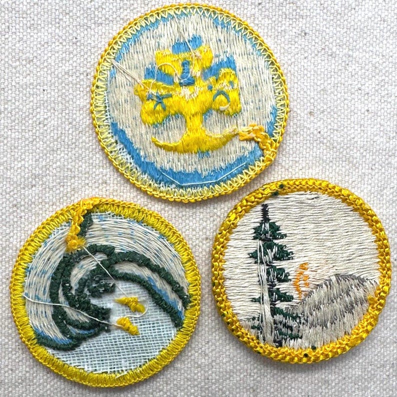 May include: Three embroidered patches with yellow, blue, and green designs. One patch features a yellow tree on a blue and white background. Another shows green waves with yellow accents. The third depicts a mountain scene with a tree.