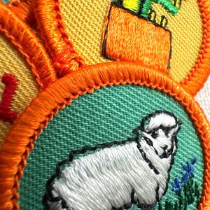 May include: Close-up of embroidered patches with orange borders. One patch features a white sheep with black outlines on a green background. Another shows a green figure on an orange box against a yellow background. The patches are likely for a youth organization.