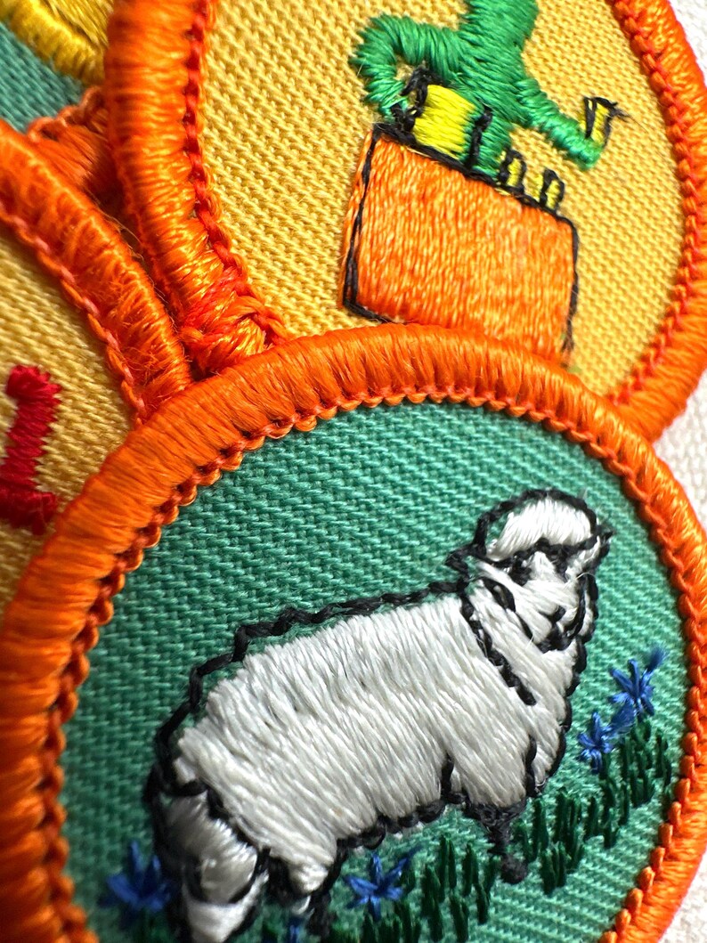 May include: Close-up of embroidered patches with orange borders. One patch features a white sheep with black outlines on a green background. Another shows a green figure on an orange box against a yellow background. The patches are likely for a youth organization.