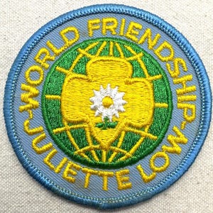 Peut inclure: &Eacute;cusson brod&eacute; avec une bordure bleu clair et les mots "WORLD FRIENDSHIP JULIETTE LOW" en jaune. Le centre pr&eacute;sente un motif jaune et vert avec une fleur blanche.