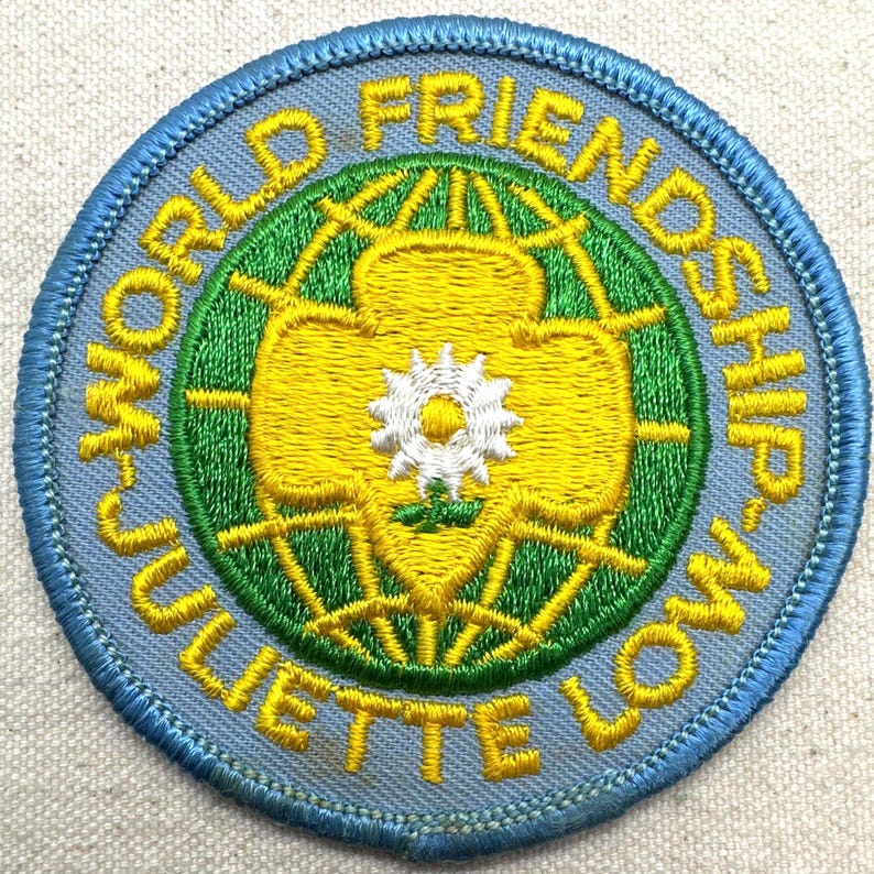Peut inclure: &Eacute;cusson brod&eacute; avec une bordure bleu clair et les mots "WORLD FRIENDSHIP JULIETTE LOW" en jaune. Le centre pr&eacute;sente un motif jaune et vert avec une fleur blanche.