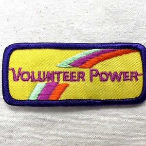May include: Embroidered patch with the words "VOLUNTEER POWER" in purple on a yellow background. The patch has a purple border and features colorful, stylized wing designs in green, purple, and orange.
