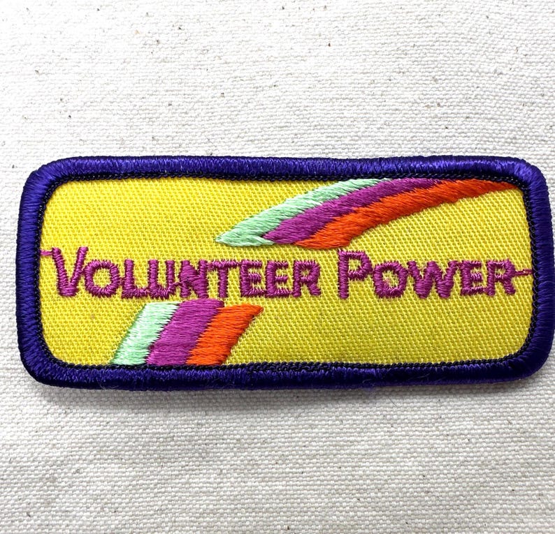 May include: Embroidered patch with the words "VOLUNTEER POWER" in purple on a yellow background. The patch has a purple border and features colorful, stylized wing designs in green, purple, and orange.