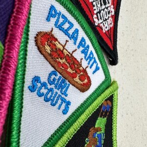 Puede incluir: Primer plano de varios parches de Girl Scouts. Un parche presenta una pizza con las palabras "Pizza Party" y "Girl Scouts". Otro parche tiene las palabras "Girl Scouts at the Theater". Los parches tienen detalles bordados y dise&ntilde;os coloridos.