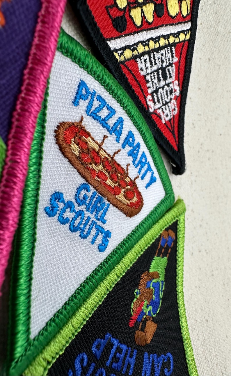 Puede incluir: Primer plano de varios parches de Girl Scouts. Un parche presenta una pizza con las palabras "Pizza Party" y "Girl Scouts". Otro parche tiene las palabras "Girl Scouts at the Theater". Los parches tienen detalles bordados y dise&ntilde;os coloridos.