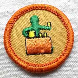 May include: A round patch with an orange border and a yellow center. The patch features an embroidered green figure holding a small object above an orange object. The design is outlined in black.