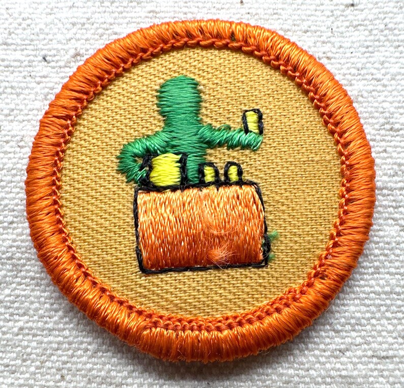 May include: A round patch with an orange border and a yellow center. The patch features an embroidered green figure holding a small object above an orange object. The design is outlined in black.