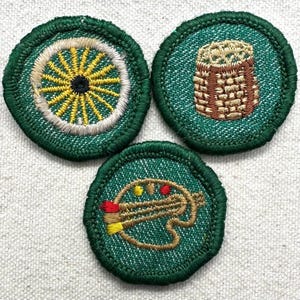 May include: Three embroidered patches with green borders. One patch features a yellow and white wheel design, another a brown basket, and the third a palette with paintbrushes and paint blobs. The patches are on a light-colored fabric.