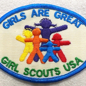 May include: Embroidered oval patch with the text "GIRLS ARE GREAT" and "GIRL SCOUTS USA". The patch features five stylized figures in yellow, orange, purple, blue, and red, with arms outstretched.