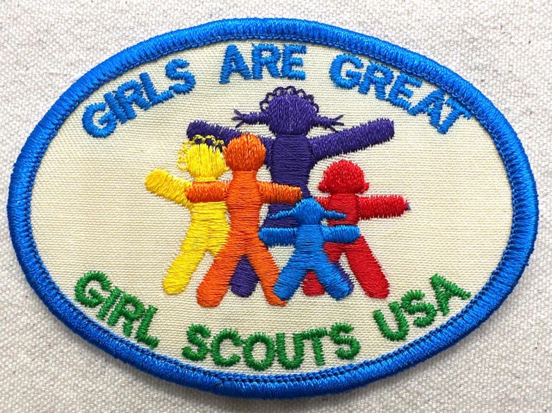May include: Oval embroidered patch with the text "GIRLS ARE GREAT" and "GIRL SCOUTS USA". The patch features five stylized figures in yellow, orange, purple, blue, and red, with arms outstretched.