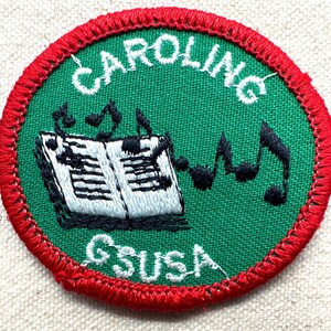 May include: Embroidered patch with a green center and red border. The patch features the word "CAROLING" in white, arched at the top, and "GSUSA" at the bottom. A musical score and notes are in the center.