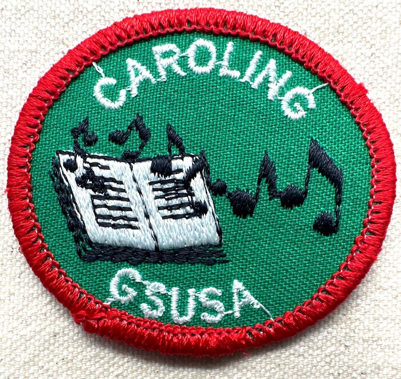May include: Embroidered patch with a green center and red border. The patch features the word "CAROLING" in white, arched at the top, and "GSUSA" at the bottom. A musical score and notes are in the center.