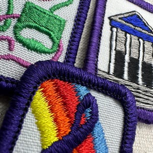 May include: Close-up of four embroidered patches with purple borders. Designs include a green and pink teacup, a building with columns, and a rainbow design. The patches are on a light-colored fabric.