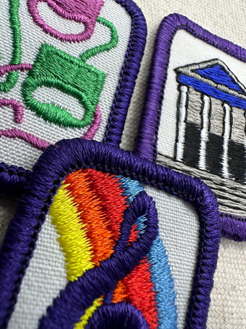 May include: Close-up of four embroidered patches with purple borders. Designs include a green and pink teacup, a building with columns, and a rainbow design. The patches are on a light-colored fabric.