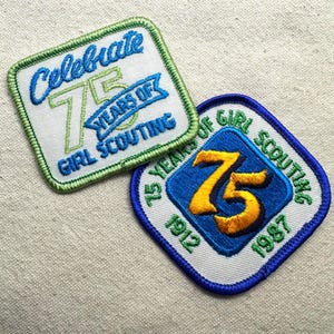 May include: Two embroidered patches celebrating 75 years of Girl Scouting. One patch features the text "Celebrate 75 Years of Girl Scouting" in blue and green. The other patch has a blue diamond with a gold "75" and the text "75 Years of Girl Scouting 1912-1987".