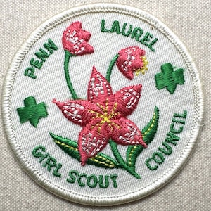 May include: Embroidered Girl Scout patch featuring a pink flower with yellow accents and green leaves. The patch is round with the words "PENN GIRL SCOUT COUNCIL" and "LAUREL" in green lettering. Two green shamrock shapes are also present.