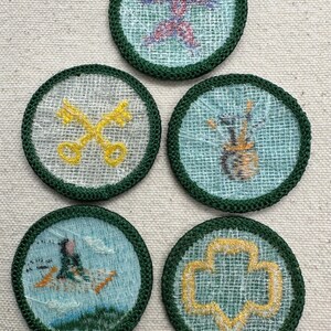 May include: Five round embroidered patches with green borders. The patches feature various designs, including a red and purple starburst, crossed yellow keys, a vase with paintbrushes, a person in a boat, and a yellow trefoil.