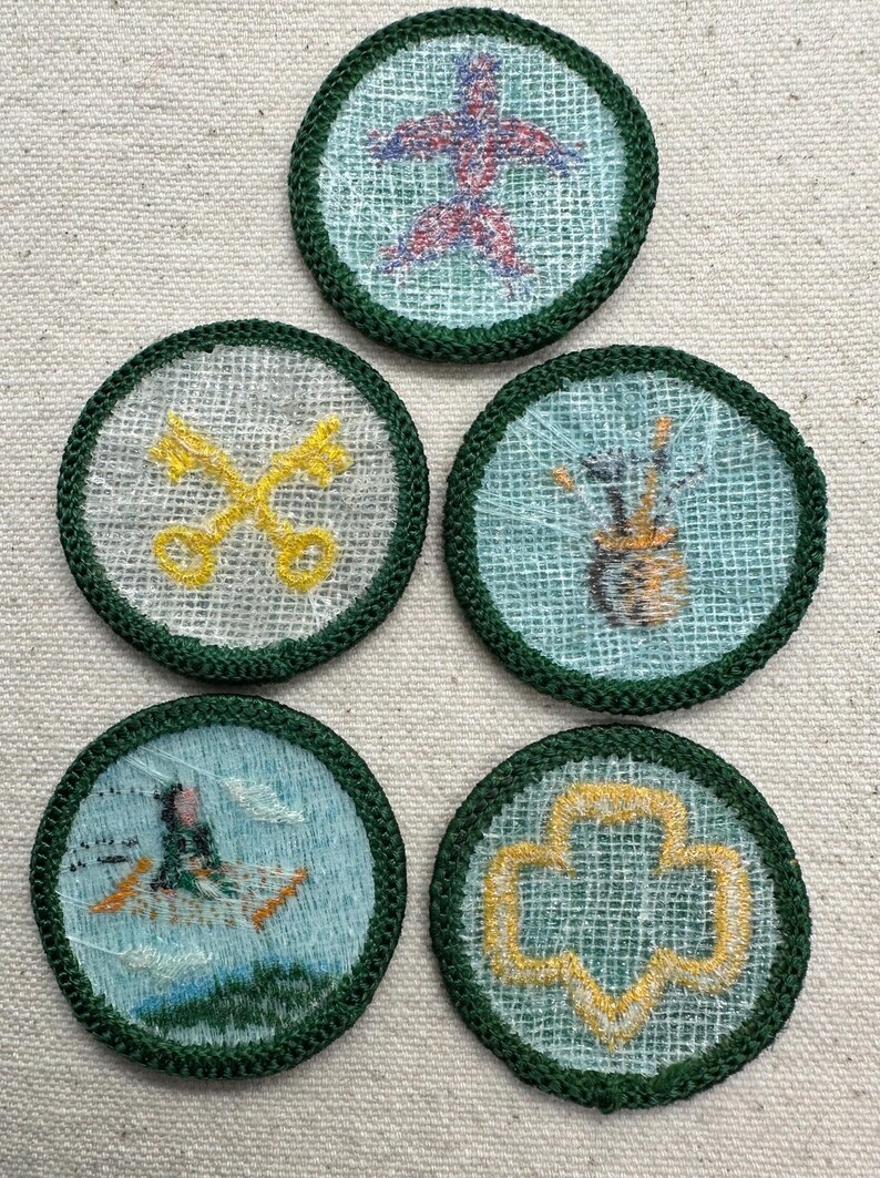 May include: Five round embroidered patches with green borders. The patches feature various designs, including a red and purple starburst, crossed yellow keys, a vase with paintbrushes, a person in a boat, and a yellow trefoil.