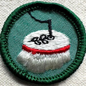 May include: Embroidered patch with a green border and a central design. The design features a white object with a red band, topped with a black element. The background is a textured green fabric.