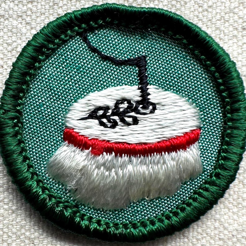 May include: Embroidered patch with a green border and a central design. The design features a white object with a red band, topped with a black element. The background is a textured green fabric.