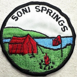 May include: Embroidered patch with a camping scene. The patch features a red tent, a blue lake, and green hills. The text "SONI SPRINGS" is arched across the top. A campfire and a red flag are also visible.