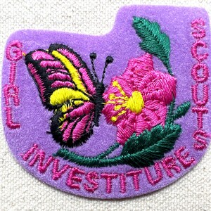 May include: A purple embroidered patch with a butterfly and flower design. The butterfly has pink, yellow, and black details. The flower is pink with a yellow center. The patch has the words "GIRL SCOUTS INVESTITURE" embroidered around the edges.