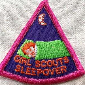 Puede incluir: Parche bordado de Girl Scouts Sleepover. El parche triangular presenta un fondo morado con una ni&ntilde;a durmiendo en una manta verde, una luna creciente y las palabras "GIRL SCOUTS SLEEPOVER" en naranja. El parche est&aacute; bordeado de rosa.