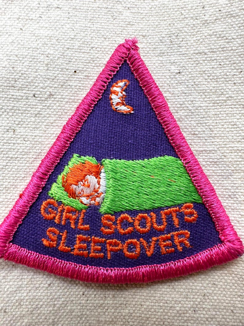 Puede incluir: Parche bordado de Girl Scouts Sleepover. El parche triangular presenta un fondo morado con una ni&ntilde;a durmiendo en una manta verde, una luna creciente y las palabras "GIRL SCOUTS SLEEPOVER" en naranja. El parche est&aacute; bordeado de rosa.