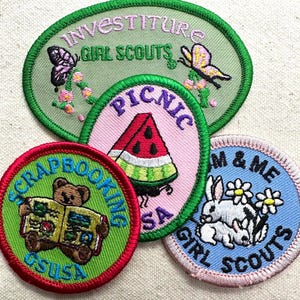 May include: A collection of Girl Scout patches with embroidered designs. The patches feature images of butterflies, a watermelon slice, a teddy bear reading, and rabbits. Text includes "Investiture", "Picnic", "Scrapbooking", and "Mom & Me".