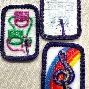 May include: Three embroidered patches with purple borders. One patch features a pink and green design, another depicts a white building, and the third showcases a rainbow and a treble clef.