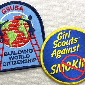 May include: Two embroidered patches. One is blue with a red ladder, a world map, and the text "BUILDING WORLD CITIZENSHIP". The other is a blue circle with "Girl Scouts Against SMOKING" and a red no-smoking symbol.