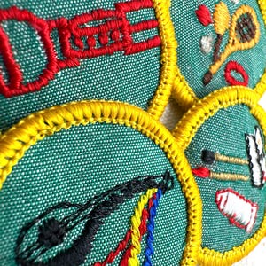 May include: Close-up of four embroidered patches with a green background and yellow trim. Each patch features a different design: a red musical instrument, a tennis racket, a black stringed instrument, and a set of paintbrushes.