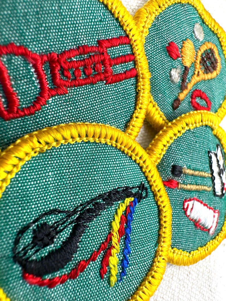 May include: Close-up of four embroidered patches with a green background and yellow trim. Each patch features a different design: a red musical instrument, a tennis racket, a black stringed instrument, and a set of paintbrushes.
