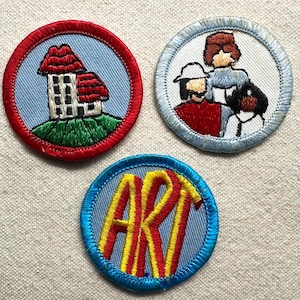 May include: Three embroidered patches. One features a house with a red roof on a green hill, another depicts two people, and the third displays the word "ART" in yellow and red on a blue background. Each patch has a circular shape.