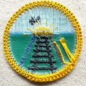 May include: Embroidered patch with a circular design featuring a black tower, yellow sunburst, and blue and green background. The patch has a yellow border and is likely for a uniform or garment.