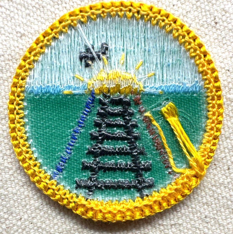 May include: Embroidered patch with a circular design featuring a black tower, yellow sunburst, and blue and green background. The patch has a yellow border and is likely for a uniform or garment.