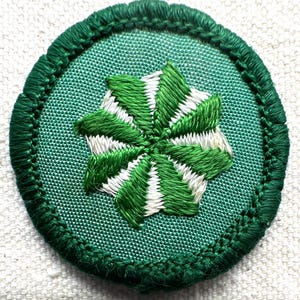 May include: A circular green embroidered patch with a central starburst design in green and white. The patch has a green border and is set against a white background. The design is detailed and appears to be hand-stitched.