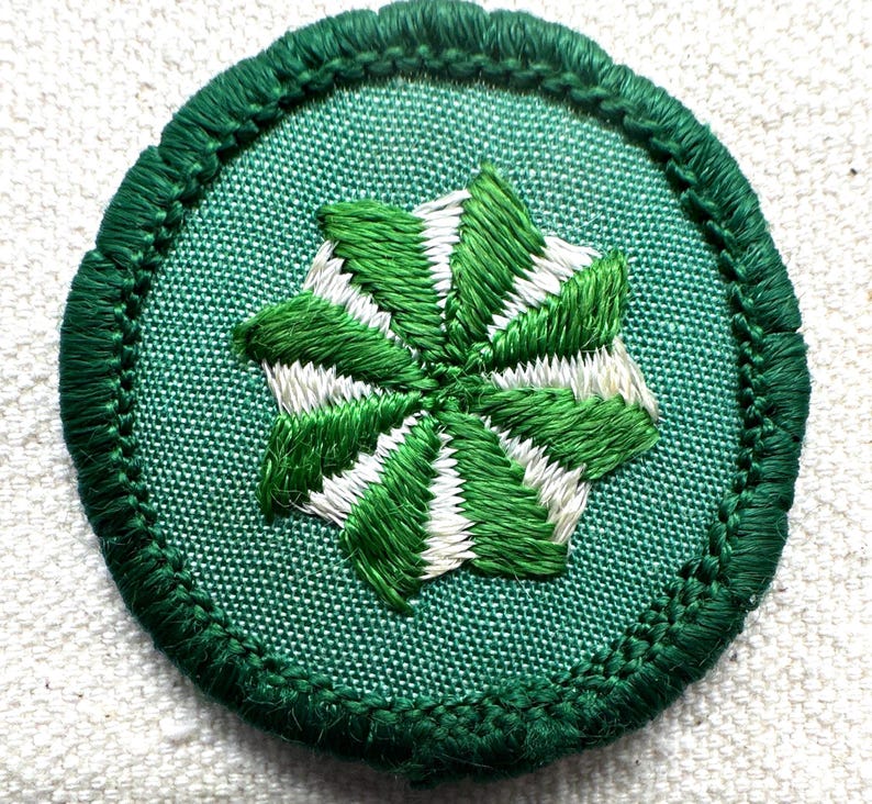 May include: A circular green embroidered patch with a central starburst design in green and white. The patch has a green border and is set against a white background. The design is detailed and appears to be hand-stitched.