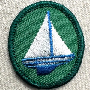 May include: Embroidered patch featuring a white sailboat with blue accents on a green circular background. The patch has a green stitched border. The sailboat design includes a blue mast and a blue hull.
