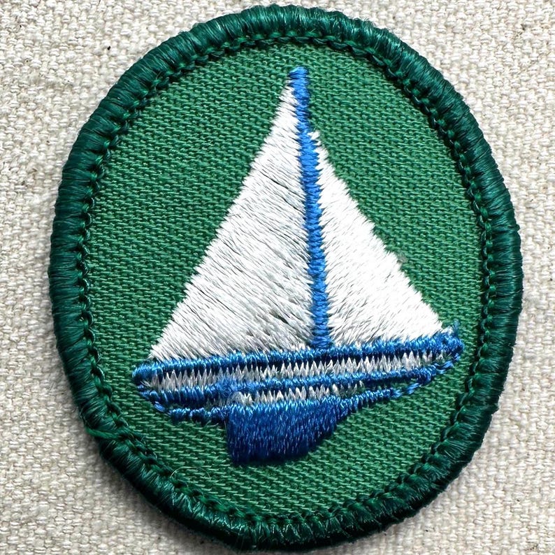 May include: Embroidered patch featuring a white sailboat with blue accents on a green circular background. The patch has a green stitched border. The sailboat design includes a blue mast and a blue hull.